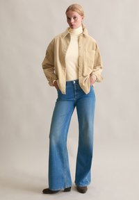 Beżowa sztruksowa koszula na jasnym golfie, zestawiona z niebieskimi, wysokim stanem, rozszerzanymi jeansami oraz brązowymi botkami na neutralnym tle.