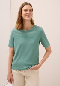 Mujer sonriendo, vistiendo una camiseta de manga corta verde claro con "ESPEJO DE PENSAMIENTOS DEL CIELO ETERNO" estampado, y pantalones beige.