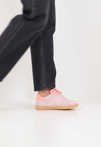 Rosa Wildledersneaker mit hellrosa Akzenten und Gummisohlen, ausgestattet mit Schnürsenkeln und diagonalen Streifen an den Seiten. Kombiniert mit schwarzer Jeans.