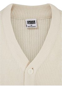 Kremo spalvos kardiganas su V formos dekolte, gaužtais audinio raštais, priekyje užsegamas mygtukais ir su etiketę, kurioje parašyta "URBAN CLASSICS, M."