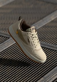 Beige sneakers med en teksturert semsket overdel, brune detaljer og gummisåle. Har snøring og en logo på tungen.