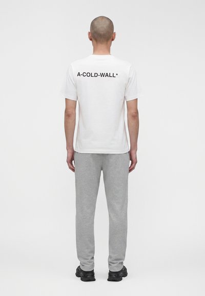 Άνδρας όρθιος με την πλάτη, φοράει λευκό T-shirt με το κείμενο «A-COLD-WALL*» στην πλάτη, γκρι φόρμες και μαύρα αθλητικά παπούτσια σε απλό λευκό φόντο.