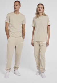Beige matching sæt bestående af en t-shirt med rund hals og joggers, begge med hvide detaljer og elastiske manchetter, lavet af blødt stof.