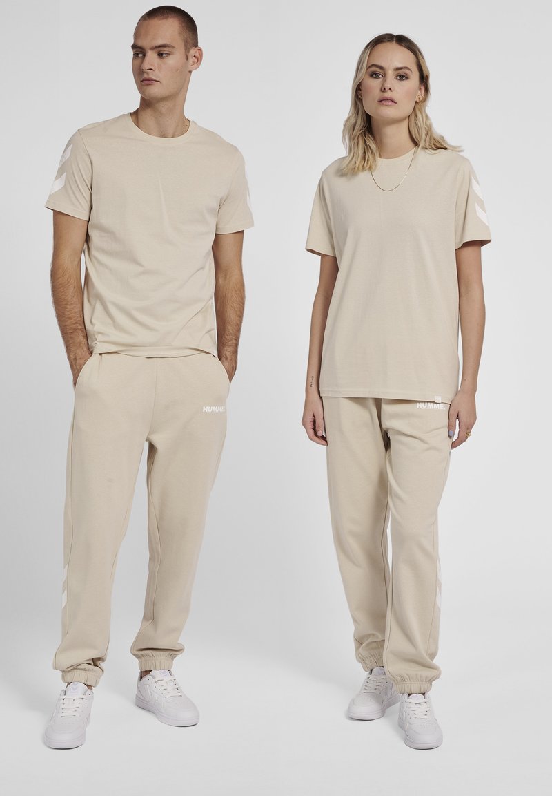 Beige matching sæt bestående af en t-shirt med rund hals og joggers, begge med hvide detaljer og elastiske manchetter, lavet af blødt stof.