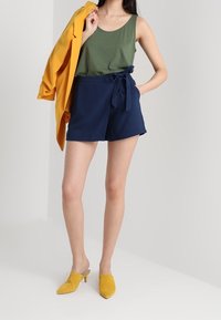 Zelený top bez rukávů, tmavě modré kraťasy s vysokým pasem a zavazovacím páskem, jasně žluté sako a žluté pantofle na podpatku na neutrálním pozadí.