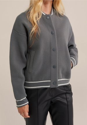 Kobieta w szarej polarowej kurtce typu varsity z białymi lamówkami i zatrzaskami, w połączeniu z czarnymi skórzanymi spodniami.