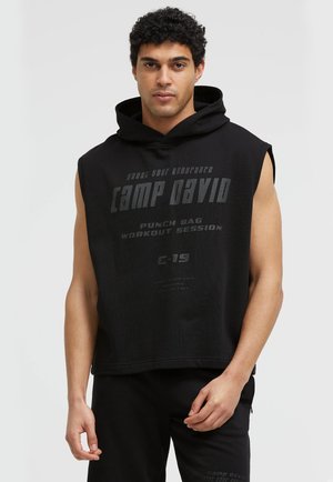 Young man with curly hair wearing a black sleeveless hoodie with text "Camp David Punch Bag Workout Session" on front.