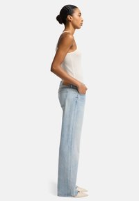 Jeans a gamba larga blu chiaro in denim con cinque tasche, un'etichetta in pelle sul retro e dettagli di cucitura sottili; abbinati a un top beige senza maniche.
