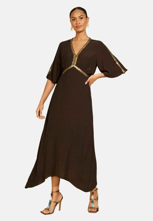 Vrouw staat en draagt een donkerbruine V-halsjurk met gouden biezen op mouwen en taille, gecombineerd met gouden sandalen met hoge hakken en kleurrijke stenen.