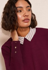 Maglione bordeaux con colletto bianco a righe, con dettagli a bottoni sulle maniche. Tessuto liscio, design attillato.