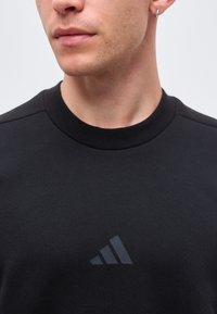 Nahaufnahme einer Person, die ein schwarzes T-Shirt mit Rundhalsausschnitt trägt, mit einem kleinen dunklen Adidas-Logo auf der Brust und einem silbernen Creolenohrring im linken Ohr.
