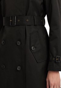 Svart trench coat med dubbelknäppta knappar, midjebälte och en sidoficka med en diagonal klaff. Slät tygtextur.