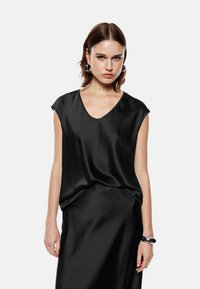 Top in satin nero con scollatura a V e maniche corte, caratterizzato da una vestibilità morbida e drappeggiata e una texture liscia. Indossato con una gonna nera coordinata.