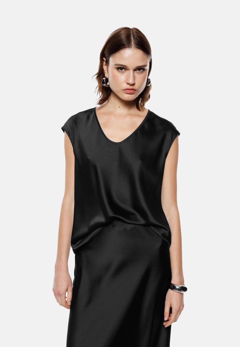 Top in satin nero con scollatura a V e maniche corte, caratterizzato da una vestibilità morbida e drappeggiata e una texture liscia. Indossato con una gonna nera coordinata.