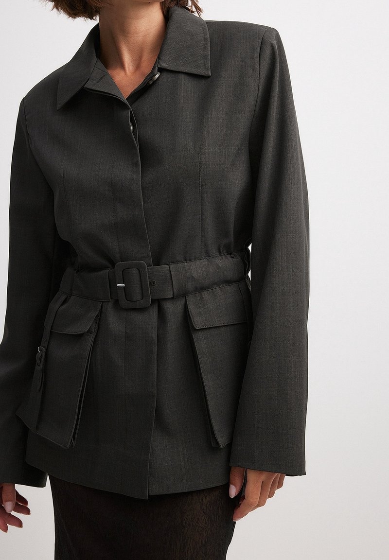 Femme portant une veste sombre ceinturée avec un large col et de grandes poches à rabat à l'avant, debout sur un fond clair uni.