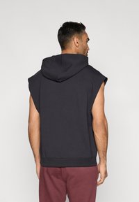 Juodos spalvos be rankovių gaubtas, pagamintas iš minkšto medvilnės audinio, su gobtuvu, ribbinių briaunų apdaila apačioje ir laisvo silueto. Galinė pusė rodo paprastą dizainą be logotipų.