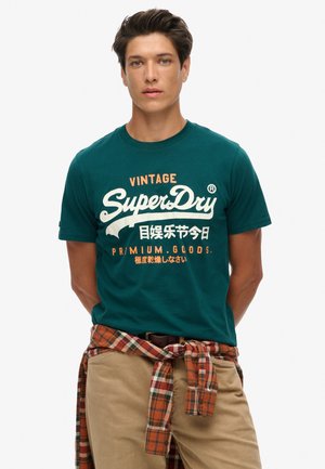 Teal bomulds t-shirt med vintage-stil "SuperDry" logo i hvid og orange. Modellen har beige bukser på med en ternet skjorte bundet om taljen.