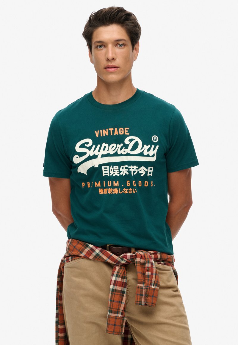 Camiseta de algodón color verde azulado con logo de estilo vintage "SuperDry" en blanco y naranja. El modelo lleva pantalones beige y una camisa a cuadros atada a la cintura.