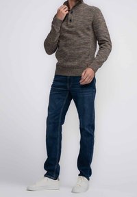 Pletený olivově hnědý svetr s vysokým límcem a dvěma černými knoflíky na výstřihu, spárovaný s modrými džíny a bílými teniskami.