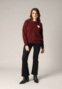 Pull en maille bourgogne avec un détail en forme de cœur blanc, associé à un jean noir évasé et des bottines à talons noires. Design simple et confortable.