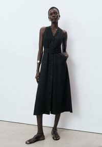 Black sleeveless dress with a button-down front and waist belt, featuring a flowing A-line silhouette. Textured fabric with pockets.