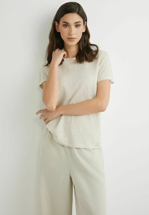 Kobieta o ciemnych włosach do ramion, ubrana w beżową koszulkę z krótkim rękawem i luźne jasne spodnie na białym tle.