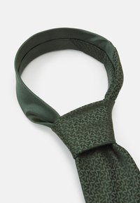 Cravate en soie vert foncé avec un motif texturé. La partie du nœud est lisse, tandis que le corps présente un design géométrique subtil.