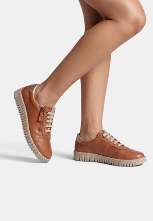 Sneakers marroni basse con lacci beige e suole in gomma indossate su gambe incrociate su sfondo bianco.
