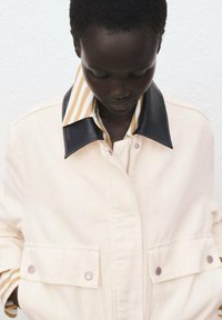 Light beige jacket with a black leather collar, featuring snap buttons and large pockets. Underneath is a striped shirt in yellow and white.