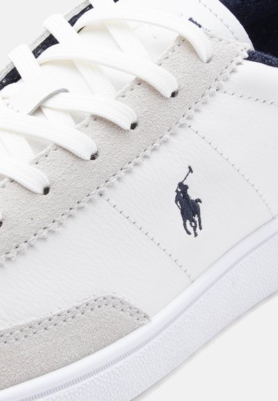 Λευκό αθλητικό παπούτσι με ανοιχτό γκρι σουέντ λεπτομέρειες, λευκά κορδόνια και μαύρο κεντητό λογότυπο παίκτη πόλο στην πλευρική επιφάνεια.