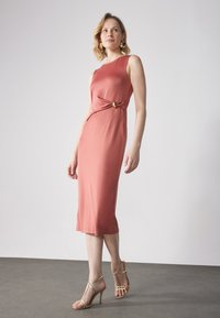 Robe ajustée, arrivant au genou, en tissu corail avec un détail de nœud torsadé à la taille, ornée d'un accent doré. Mannequin portant des talons à lanières.