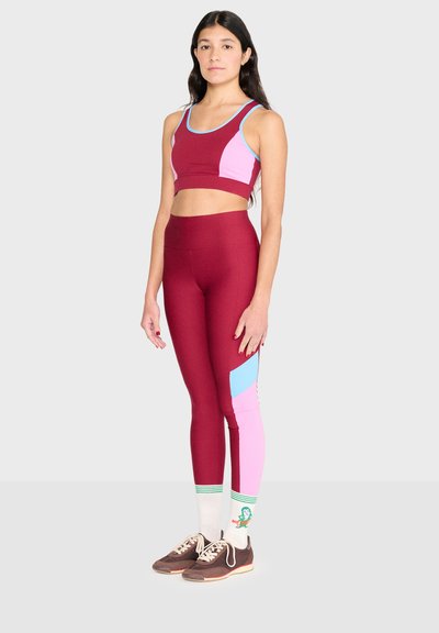 Femme debout, tournée vers l'avant, portant des leggings de sport bordeaux avec des panneaux roses et bleus, un crop top assorti, des chaussettes blanches et des baskets marron.