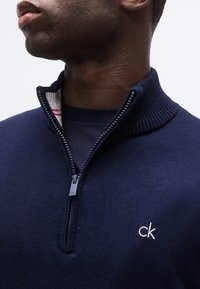 Homme portant un pull à fermeture éclair quart marine avec col côtelé, logo "ck" de Calvin Klein sur la poitrine et doublure intérieure du col rayée rouge.