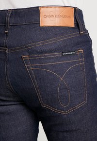 Tummansiniset farkut, joissa oranssit ompeleet, takapuolen vyötäröllä nahkainen merkki, jossa lukee "CALVIN KLEIN JEANS", ja pieni lappu taskussa.