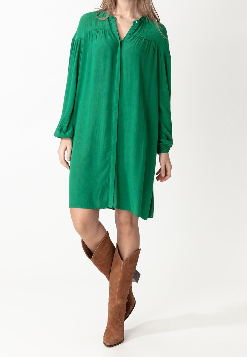 Femme portant une robe ample verte à manches longues avec des détails de boutons, associée à des bottes en daim marron mi-mollet, debout devant un fond blanc.