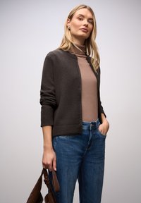 Tmavě hnědý svetr na zapínání s texturovaným povrchem, nošený přes světle hnědý rolák. Doplněn volnými modrými džínami a drží hnědou kabelku.