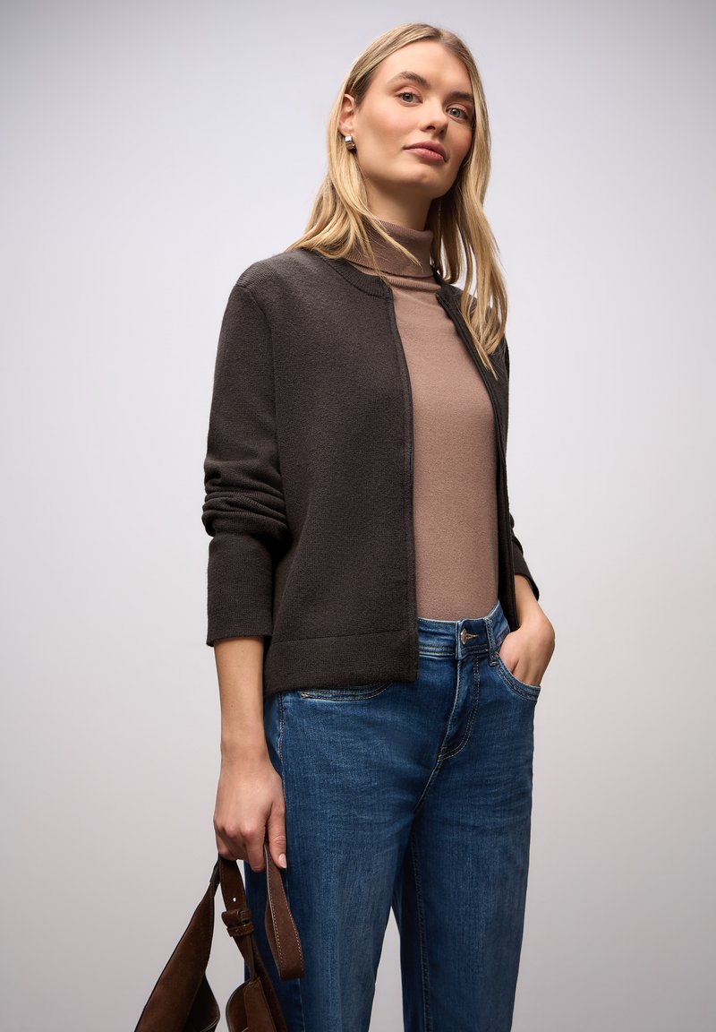 Tmavě hnědý svetr na zapínání s texturovaným povrchem, nošený přes světle hnědý rolák. Doplněn volnými modrými džínami a drží hnědou kabelku.