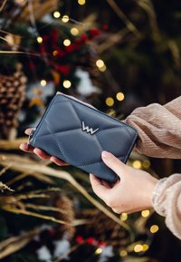 Blue leather wallet with quilted design, featuring a silver logo accent and zipper closure. Hands holding the wallet against a blurred festive background.