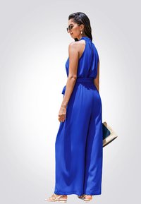 Mono sin mangas azul con cintura ajustada y pantalones anchos. Tela suave, escote plisado en la espalda, combinado con sandalias planas beige y un bolso de paja.