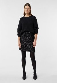 Schwarzer Strickpullover mit lockeren Ärmeln, kombiniert mit einem enganliegenden schwarzen Paillettenrock. Das Model trägt schwarze Strumpfhosen und spitze High Heels. Schlichter Hintergrund.