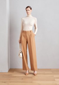 Beige gestrickter Rollkragenpullover mit gerippter Textur, kombiniert mit hochtaillierten, weit geschnittenen, camel-farbenen Hosen und nudefarbenen Absätzen. Weiße Handtasche.