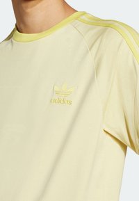 Ljusgul Adidas T-shirt med broderad trefoil-logotyp och tre ränder på axeln, bärs av en person.