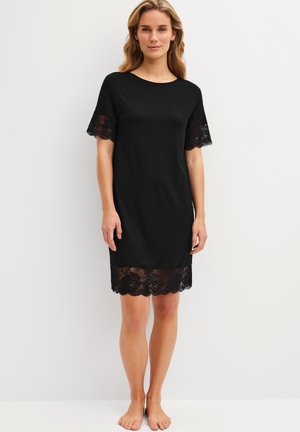 Femme portant une courte robe noire avec un bord en dentelle aux manches et à l'ourlet, debout pieds nus devant un fond clair uni.
