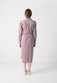 Vestito a camicia a righe rosso e bianco con maniche lunghe, colletto, vita accentuata da una cintura e lunghezza fino al polpaccio. Indossato con scarpe di colore chiaro.