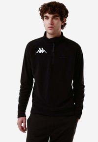 Pullover in pile nero con zip a metà, dotato di collo alto e logo bianco sul lato sinistro del petto. Texture morbida e maniche lunghe.