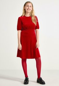 Czerwona sukienka z weluru z krótkimi bufkami, marszczoną talią i białymi kropkami; w połączeniu z czerwonymi rajstopami i czarnymi butami na platformie.