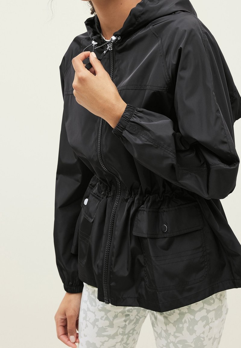 Person adjusting the zipper of a black hooded jacket with elastic cuffs and flap pockets, wearing white floral patterned pants.