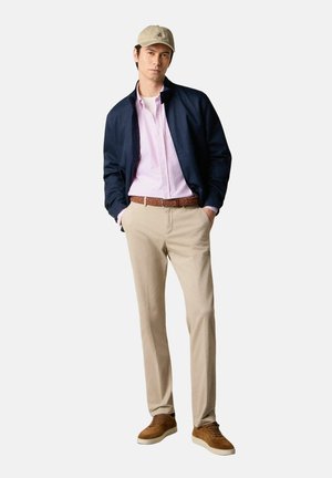 Homme portant une casquette de baseball beige, une veste bleu marine, une chemise à rayures roses, un pantalon beige, une ceinture marron et des baskets en daim marron, debout les mains dans les poches.