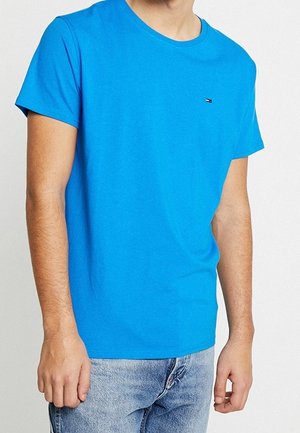 Homme portant un t-shirt uni bleu vif à manches courtes et un jean bleu clair sur un fond neutre.