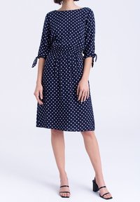 Kvinna i en marinblå knälång klänning med vita prickar och trekvartsärmar som är knutna vid armbågarna, tillsammans med svarta klacksandaler.